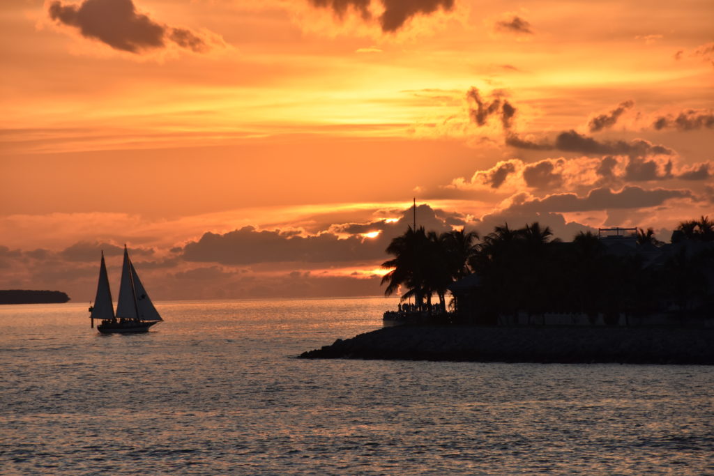 the sun is setting behind a island with a sail boat sailing towards the sinking sun with a very vibrant orange and yellow sky.