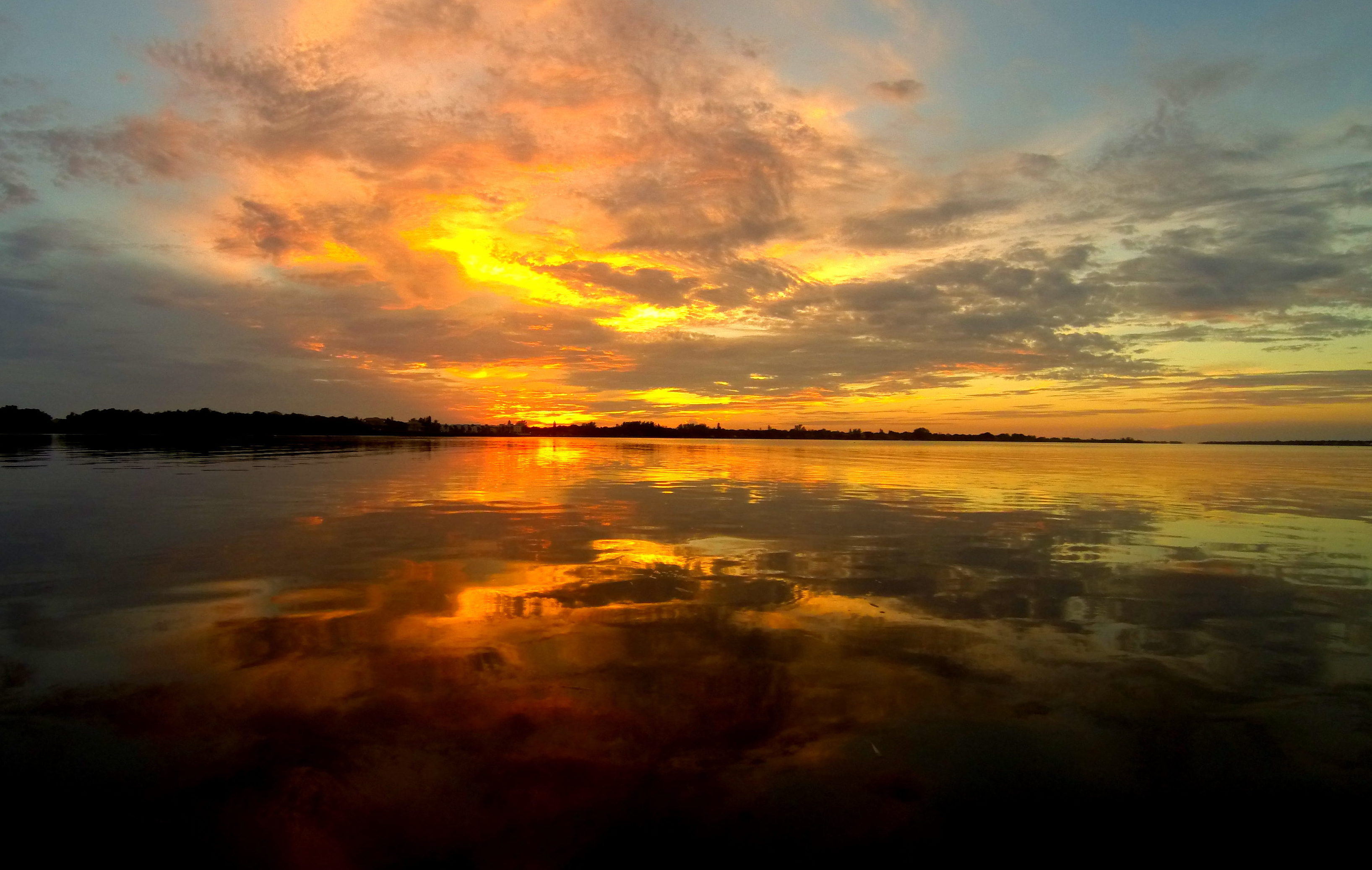 radiating orange and yellow clouds fill the sky and reflect off of the water as the as the sun sets.