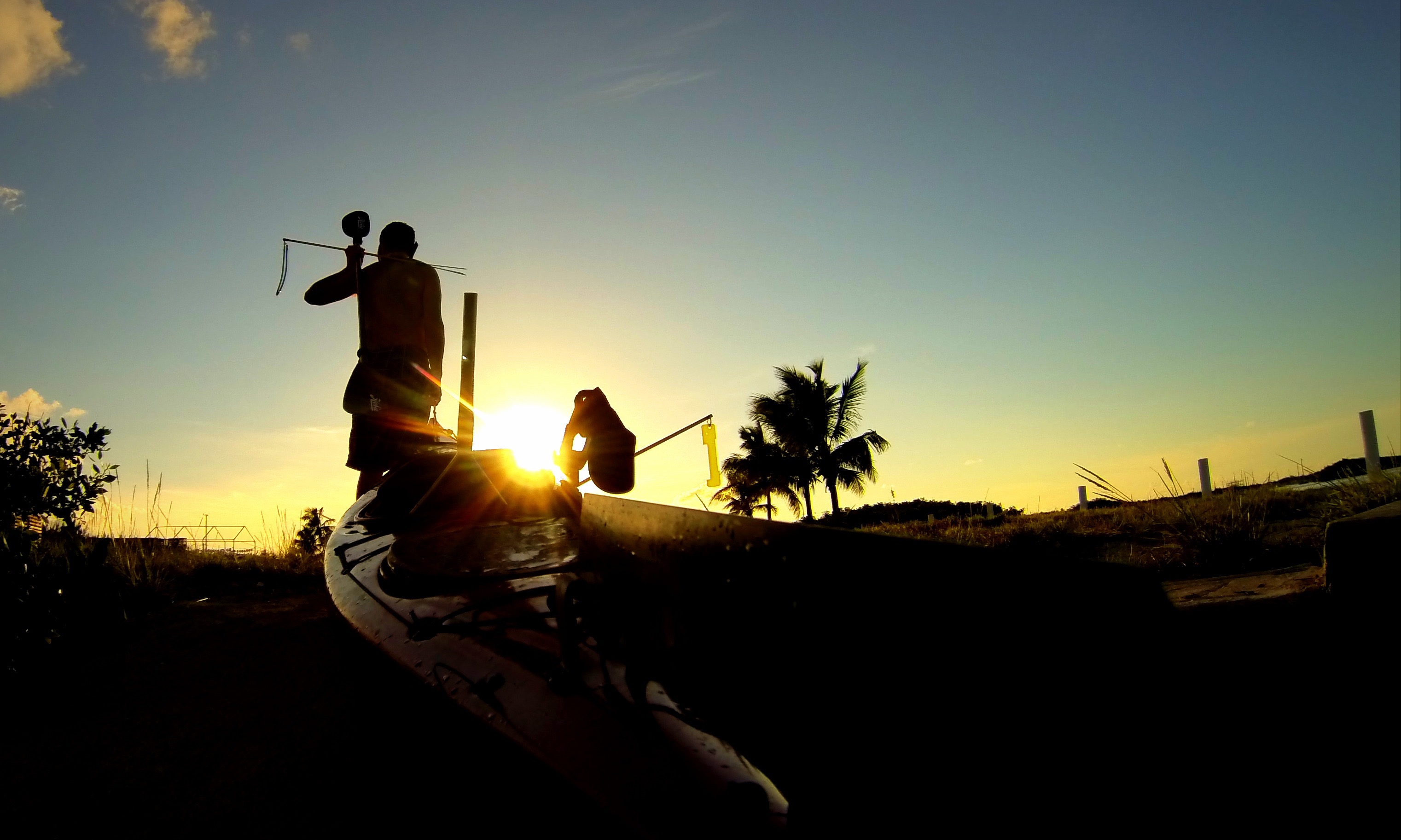 A kayak is being dragged into the sunset while someone holds a pole spear over their shoulder.